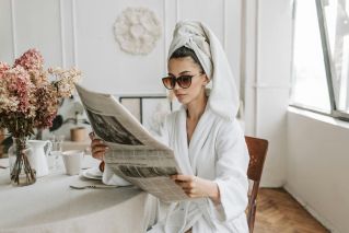 Woman in bathrobe reading a newspaper at a stylish indoor setting with flowers.