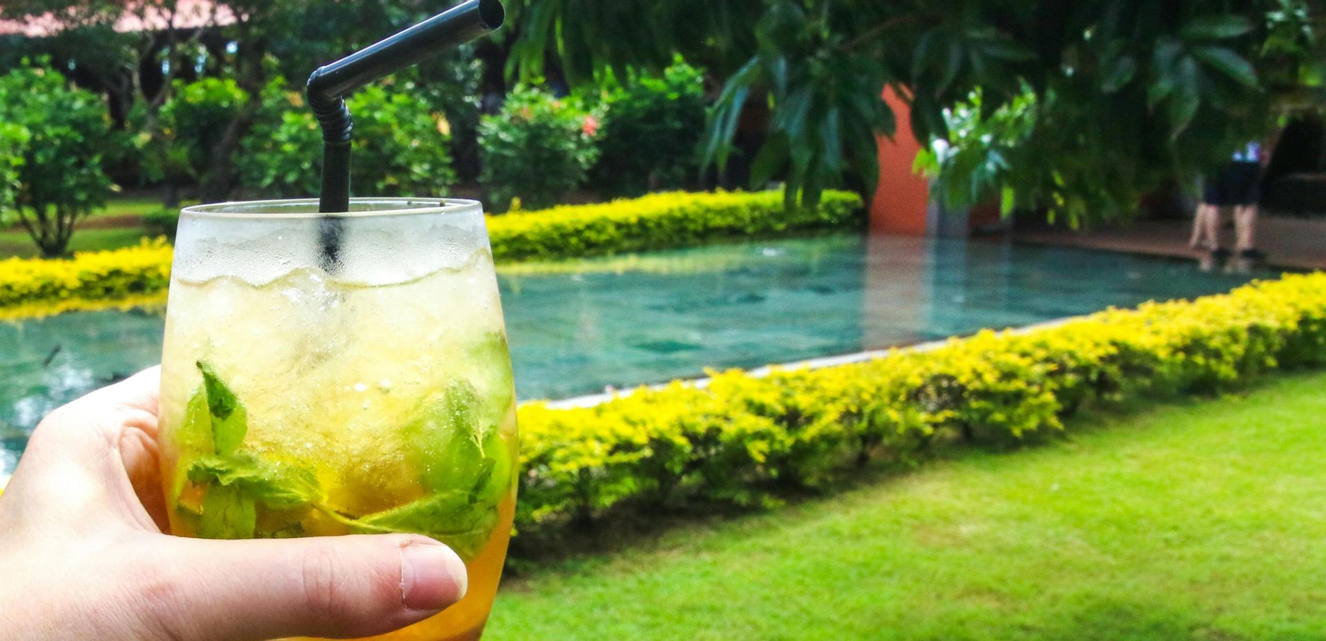 A hand holding an icy tea mocktail in a lush garden with a pool view, ideal for relaxation.