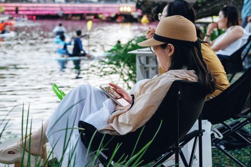 Woman lounging by a lake, watching kayakers pass by. Peaceful outdoor scene.