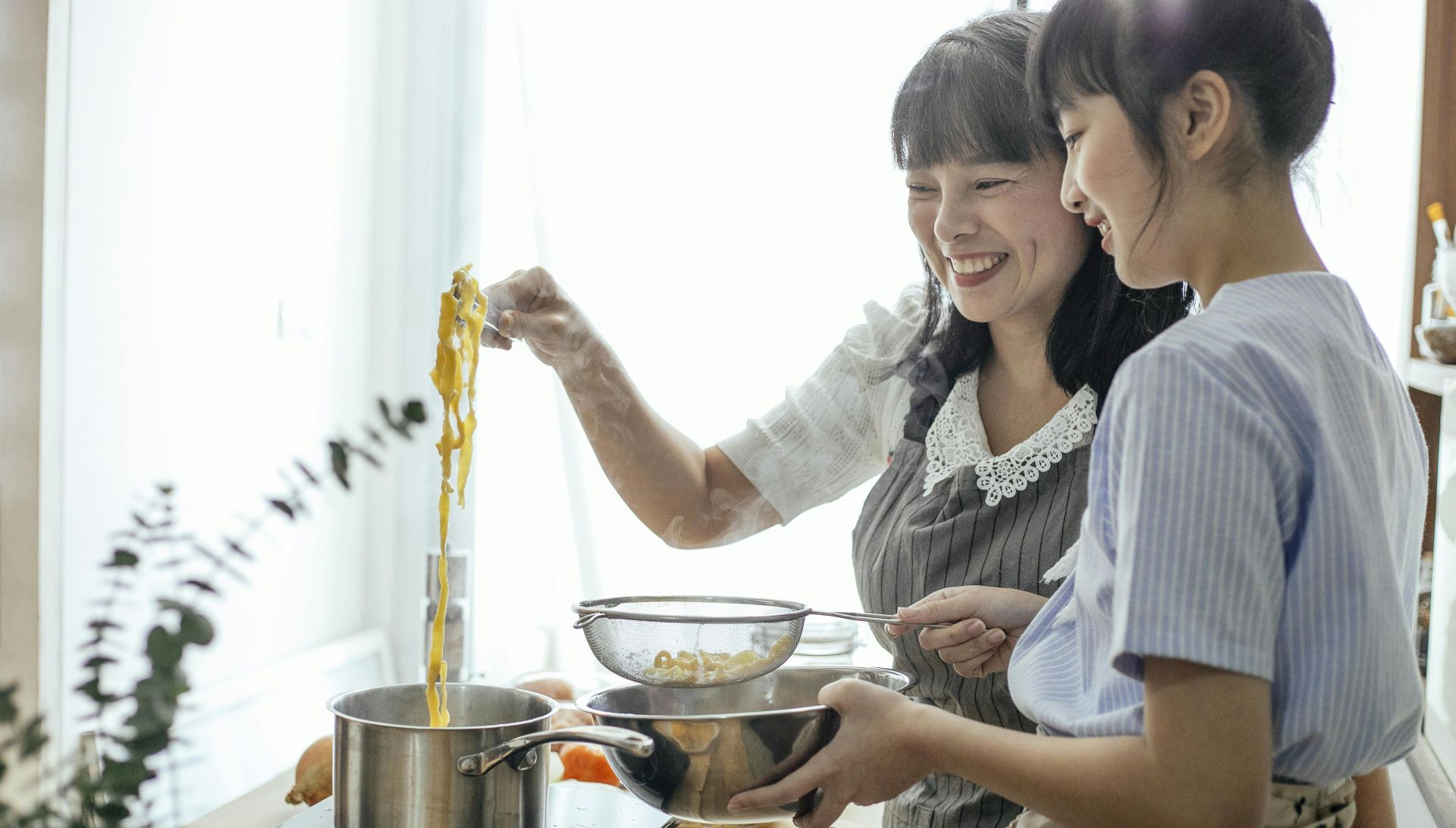Side view of smiling Asian teenager with middle age mother serving hot boiled pasta in drainer